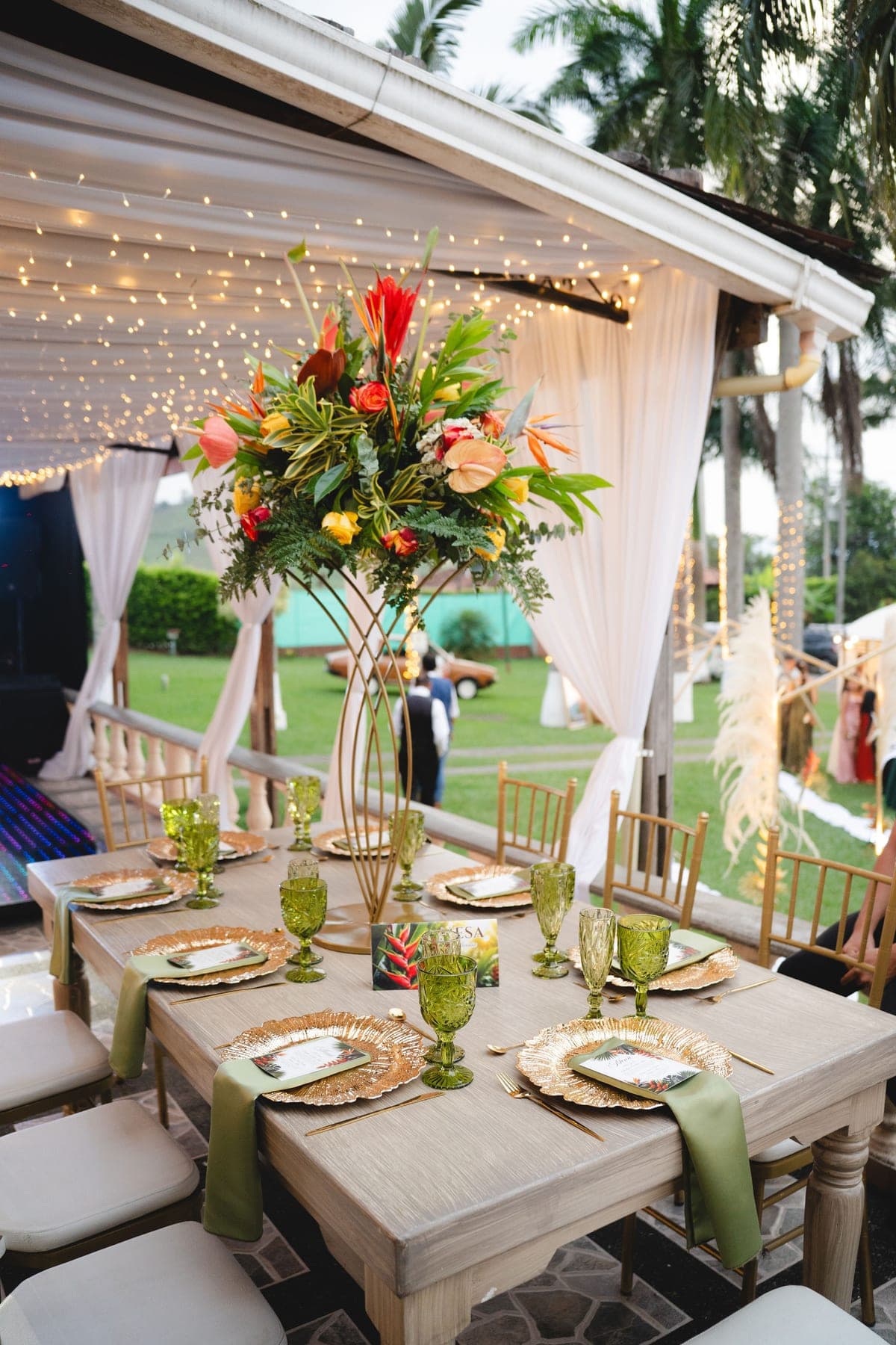 Mesa de invitados con flores tropicales y copas verdes bajo una carpa