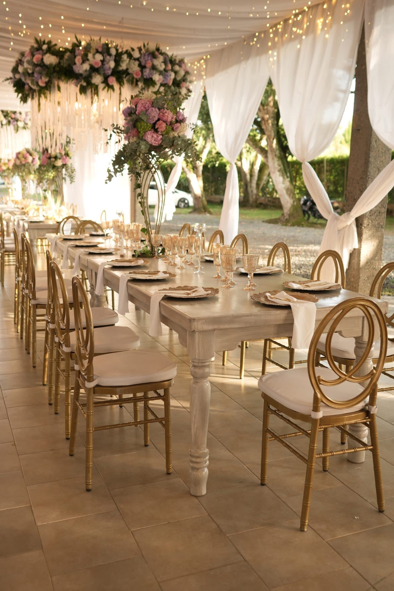 Salón de 15 años con mesas largas, sillas doradas y flores colgantes
